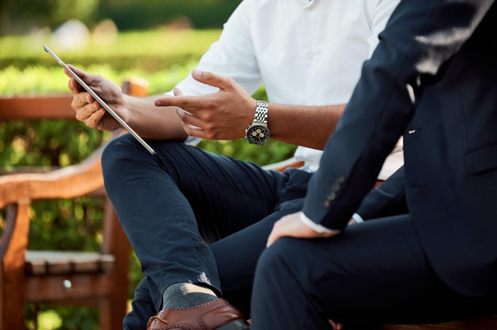 Two men sitting at park bench conducting business negotiating a real estate deal while dressed in business attire and looking at a computer screen